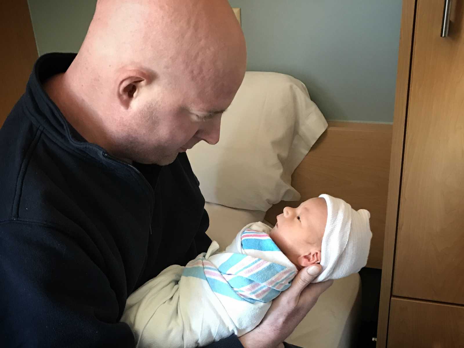 brain cancer patient holds his newborn son who is swaddled in blanket and wearing white hat