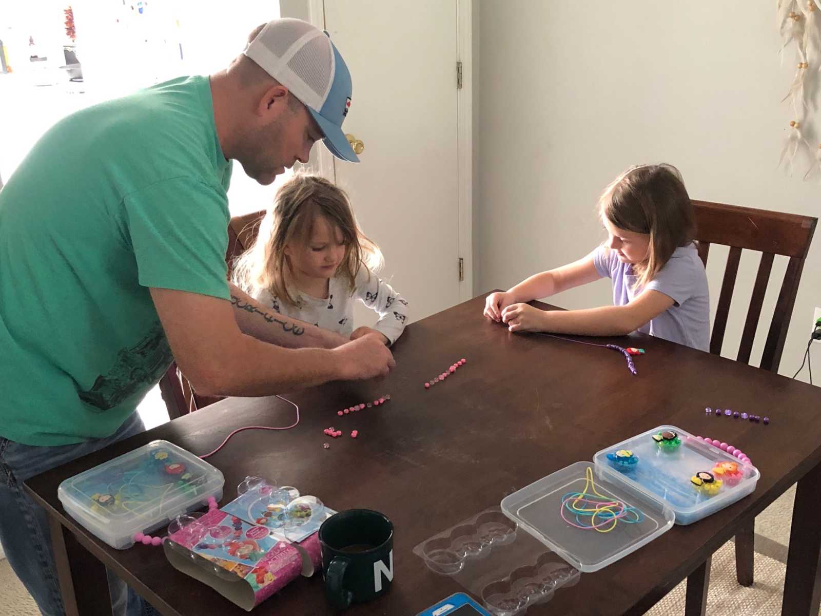 man leans over table to help stepdaughter make a bracelet while other stepdaughter sits across table making her own bracelet