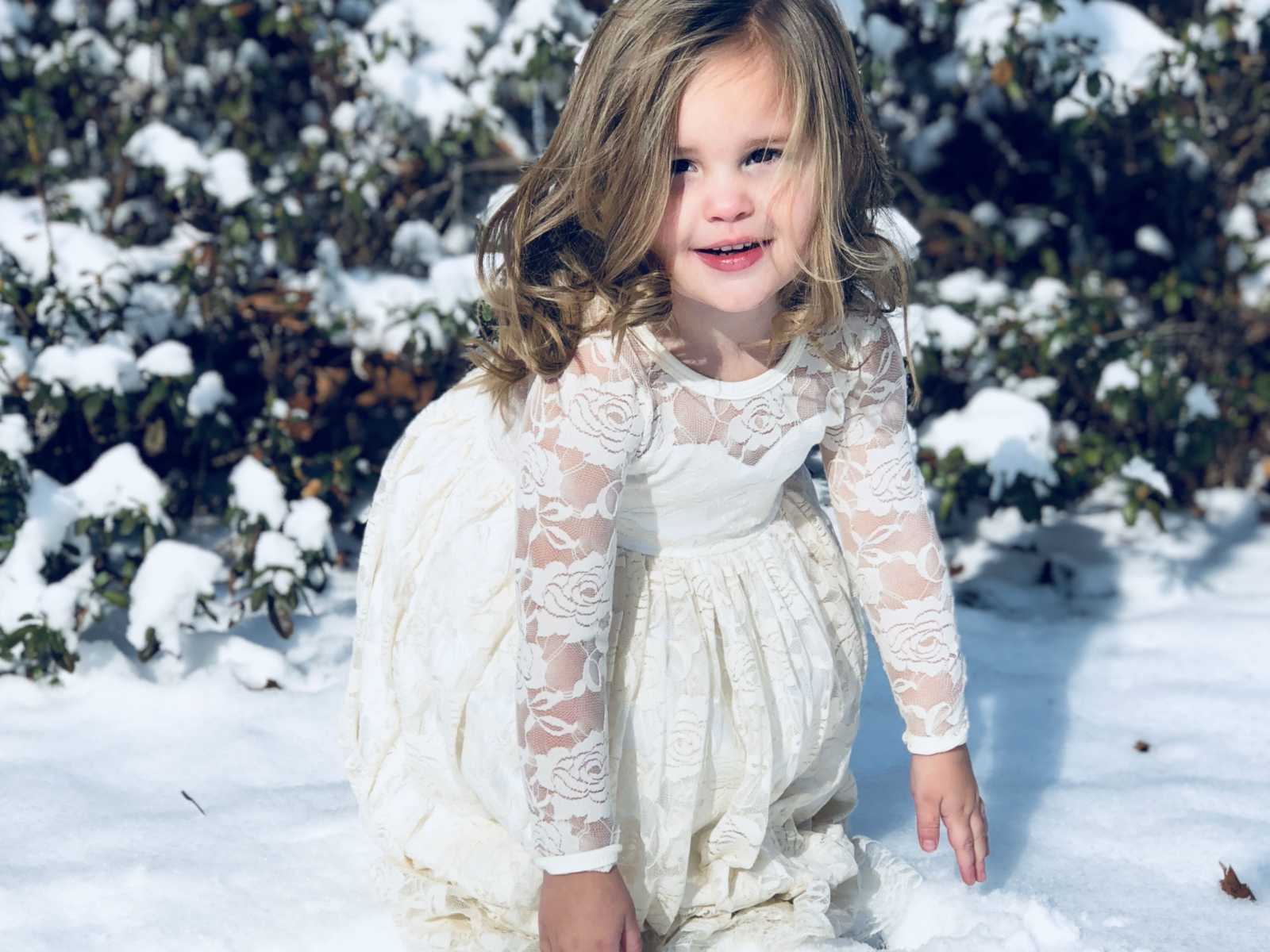young girl in white floral lace dress crouching down to the snowy ground