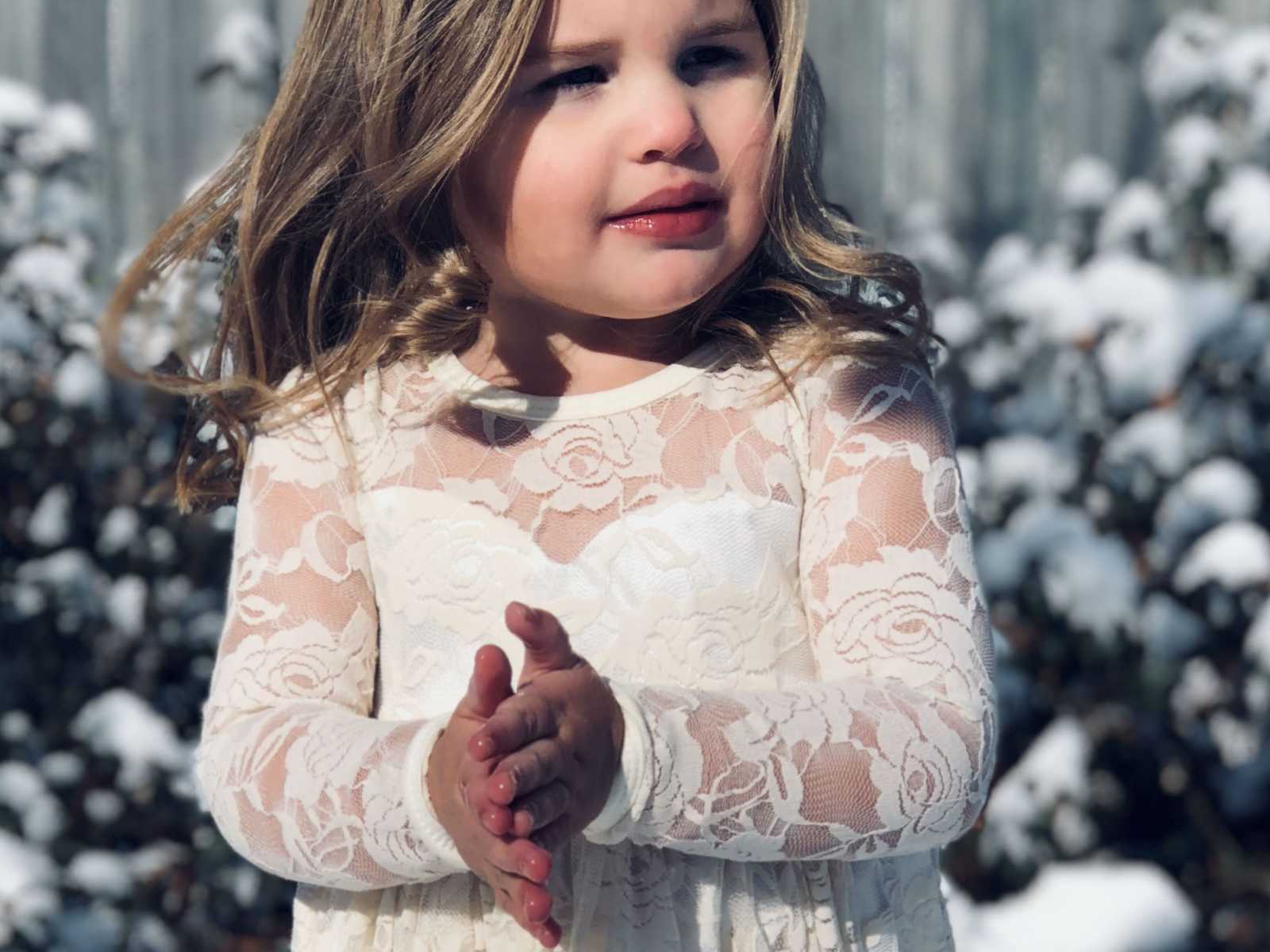 young girl in white floral lace dress looks to the side with hands together 