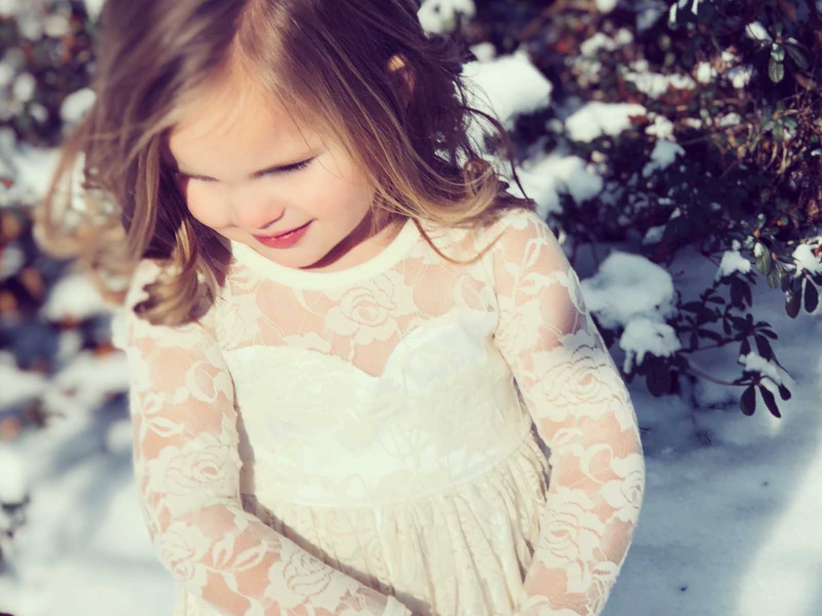 young girl in white floral lace dress looks down in front of snowy bush