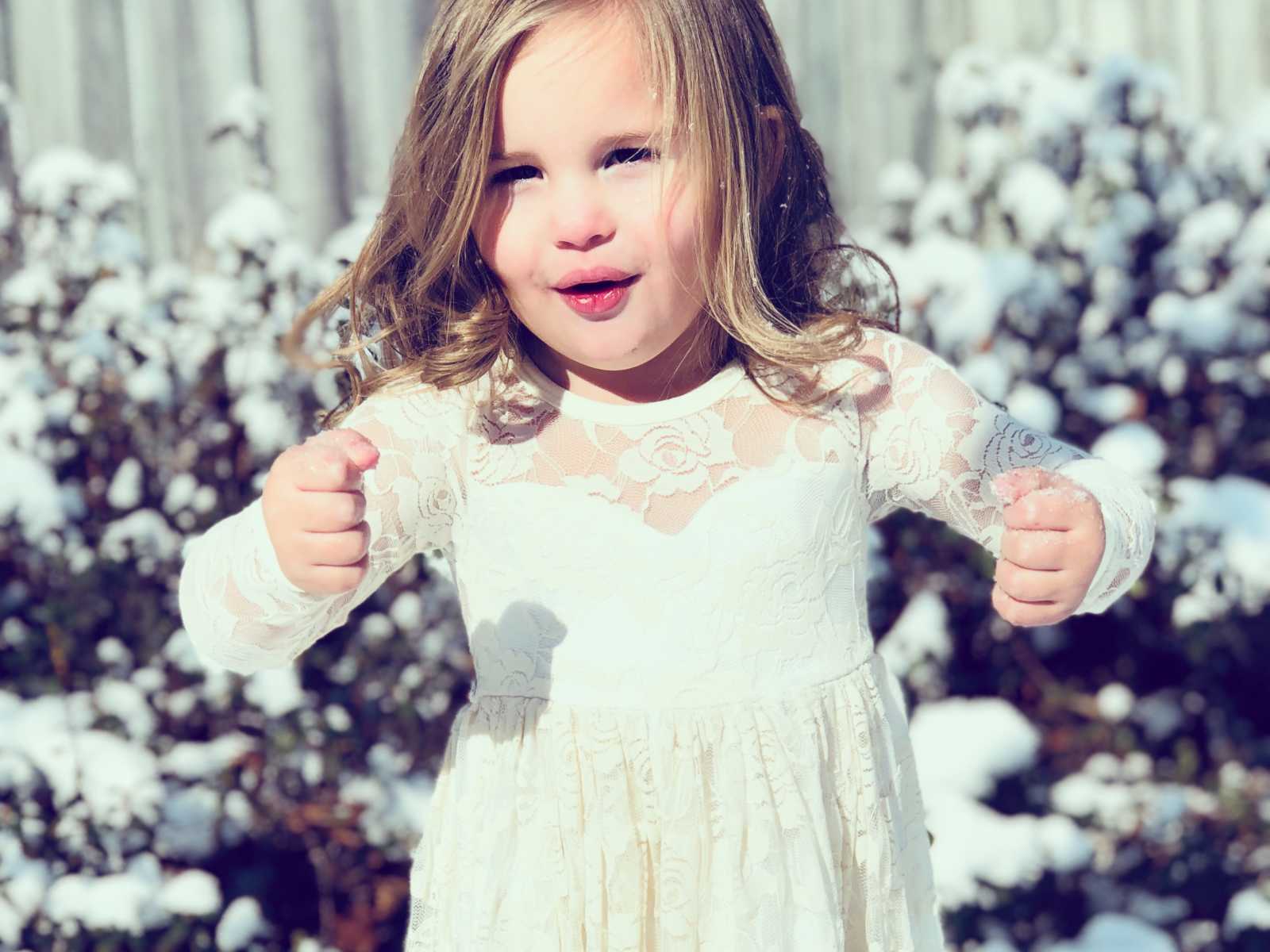 little girl in white dress with arms held up and hands in fists in front of snowy plant