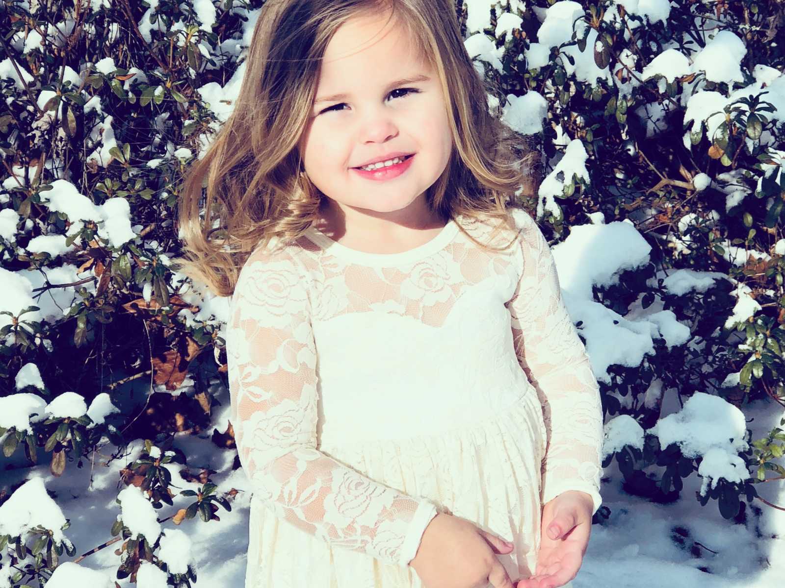young girl in white floral lace dress smiling in front of snowy bush