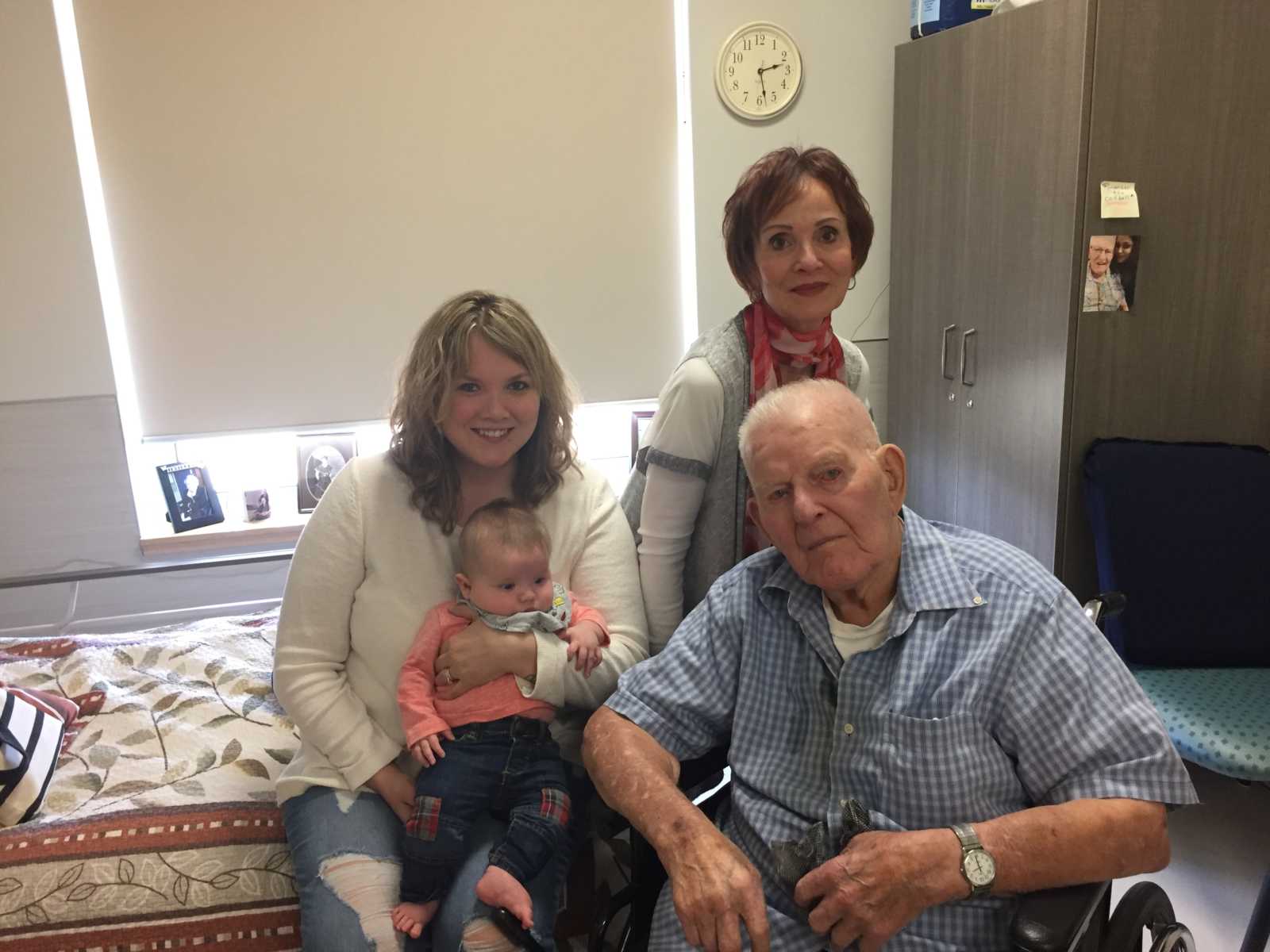 Woman sits on bed with baby in lap next to her grandma and great-grandfather is sitting in wheelchair next to bed
