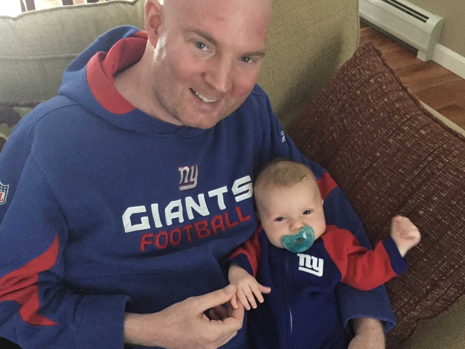 brain cancer patient in new york giants sweatshirt sits on couch with son in his lap with giants onesie on