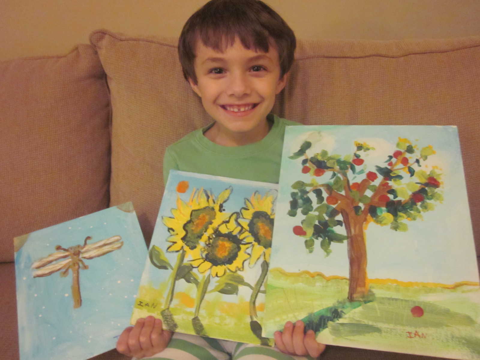 young boy sits on couch smiling while holding up three drawings of firefly, sunflower, and tree