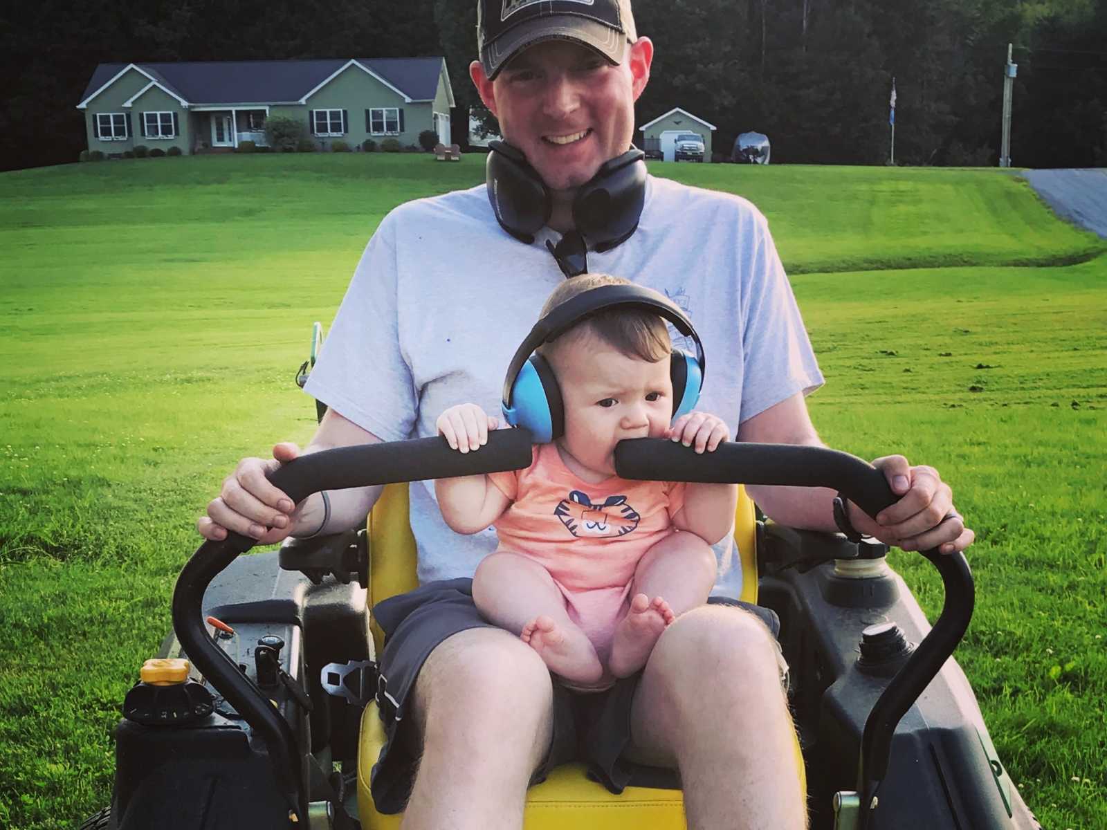 brain cancer patient sitting on lawn mower with baby in lap with headphones on with house in background