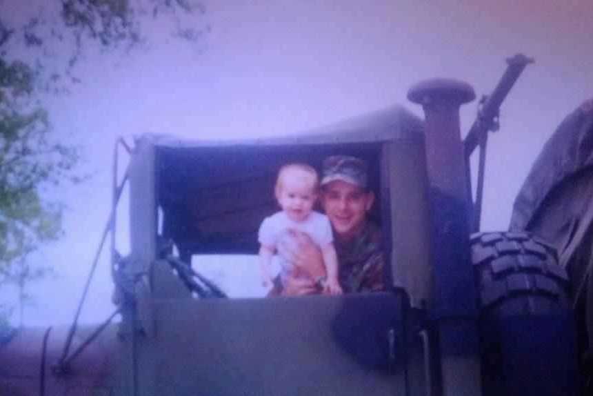soldier smiling in drivers seat of army vehicle holding infant daughter