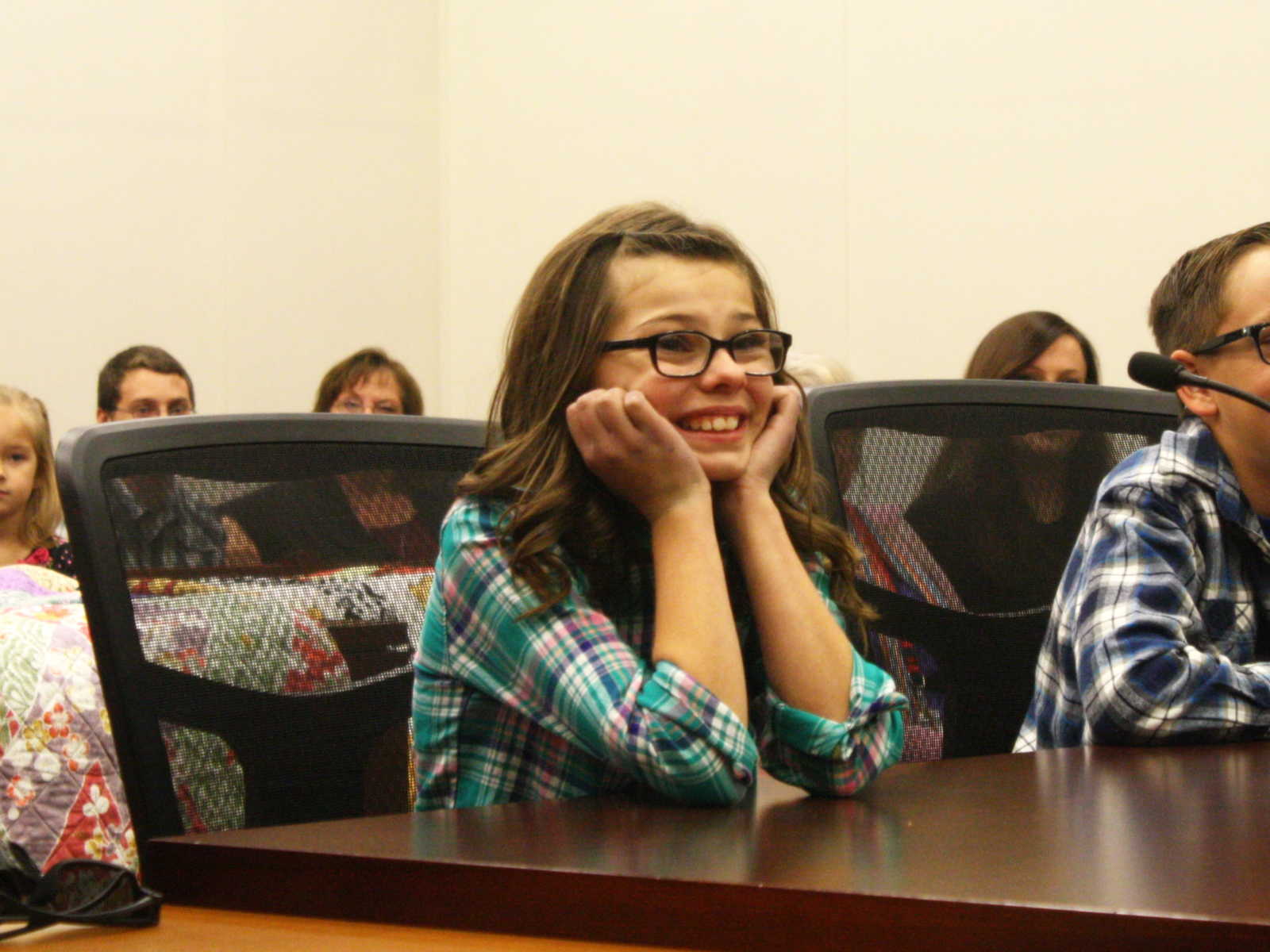 young girl with glasses smile with hands rested on chin at adoption court with brother sitting next to her 