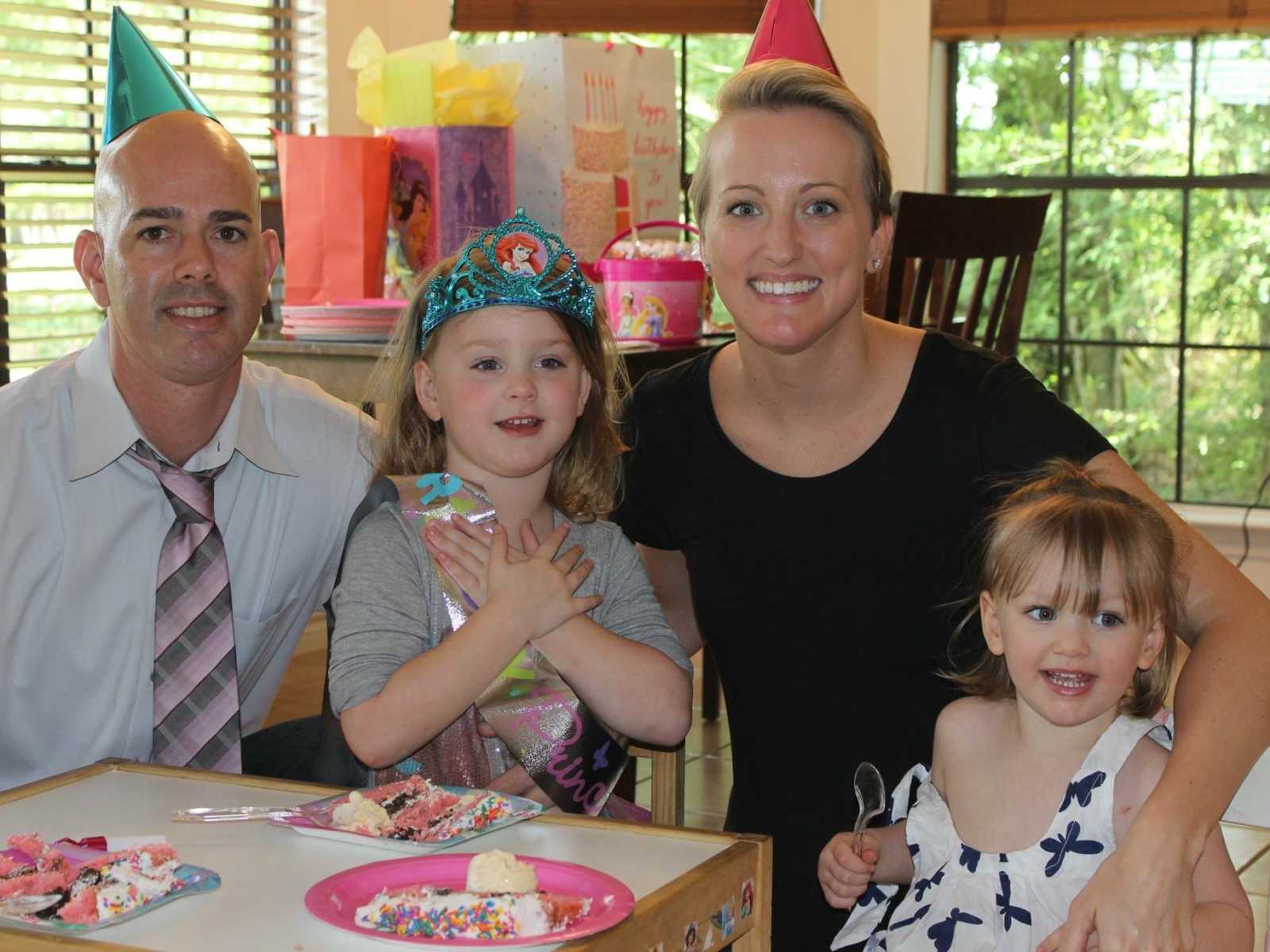 Mother and father with party hats on sit and smile with two daughter at table with cake and table of presents in background