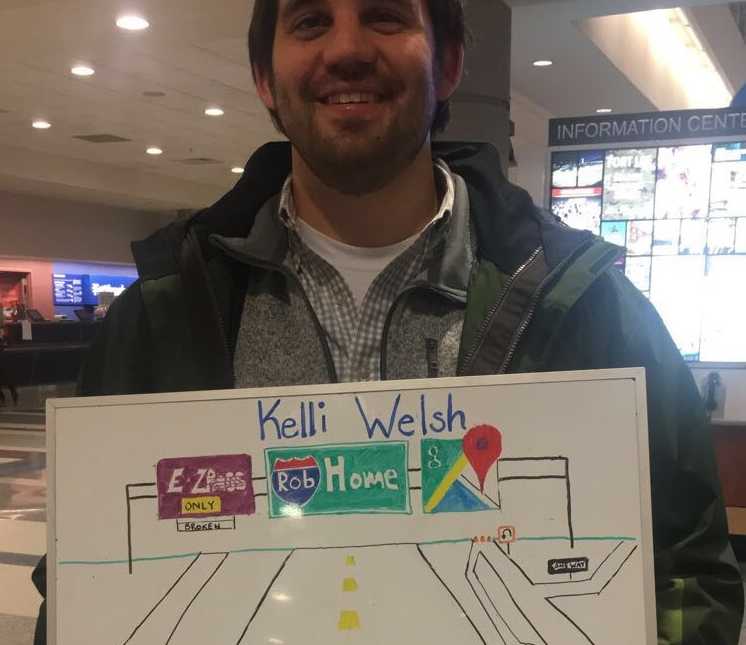 boyfriend picks up girlfriend at airport with sign mimicking an exit sign saying, “Home” with his name on it