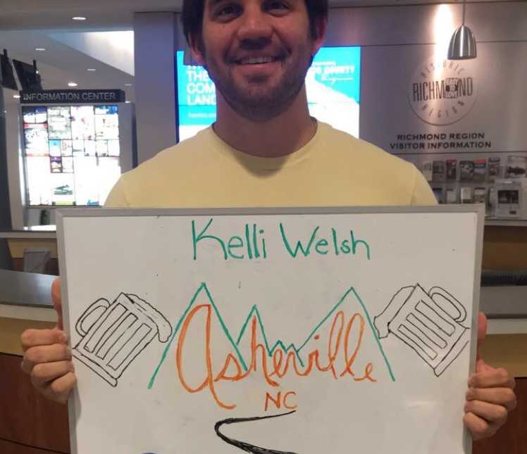 girlfriend is picked up by boyfriend at airport with sign saying, "Asheville, NC" and drawing of beer mugs