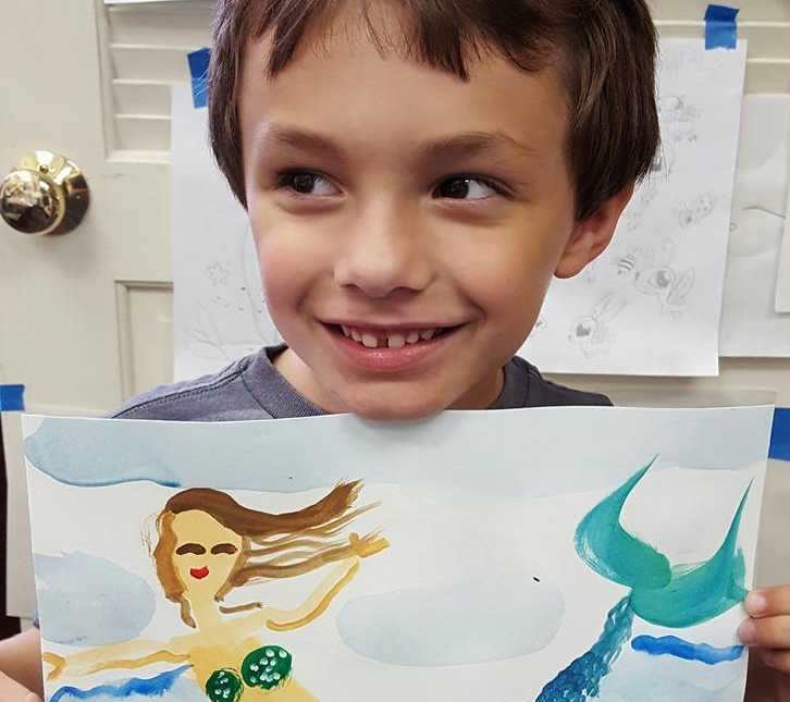 toddler boy holds up drawing of mermaid under his chin while smiling