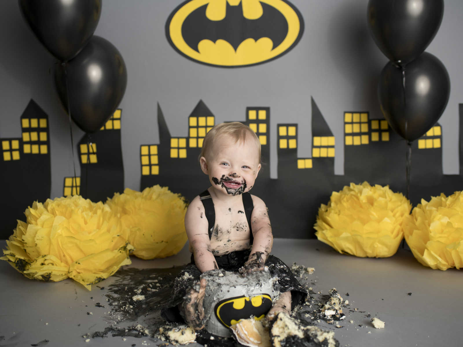 infant boy in suspenders destroying batman cake in front of backdrop of Gotham city, black balloons and batman logo