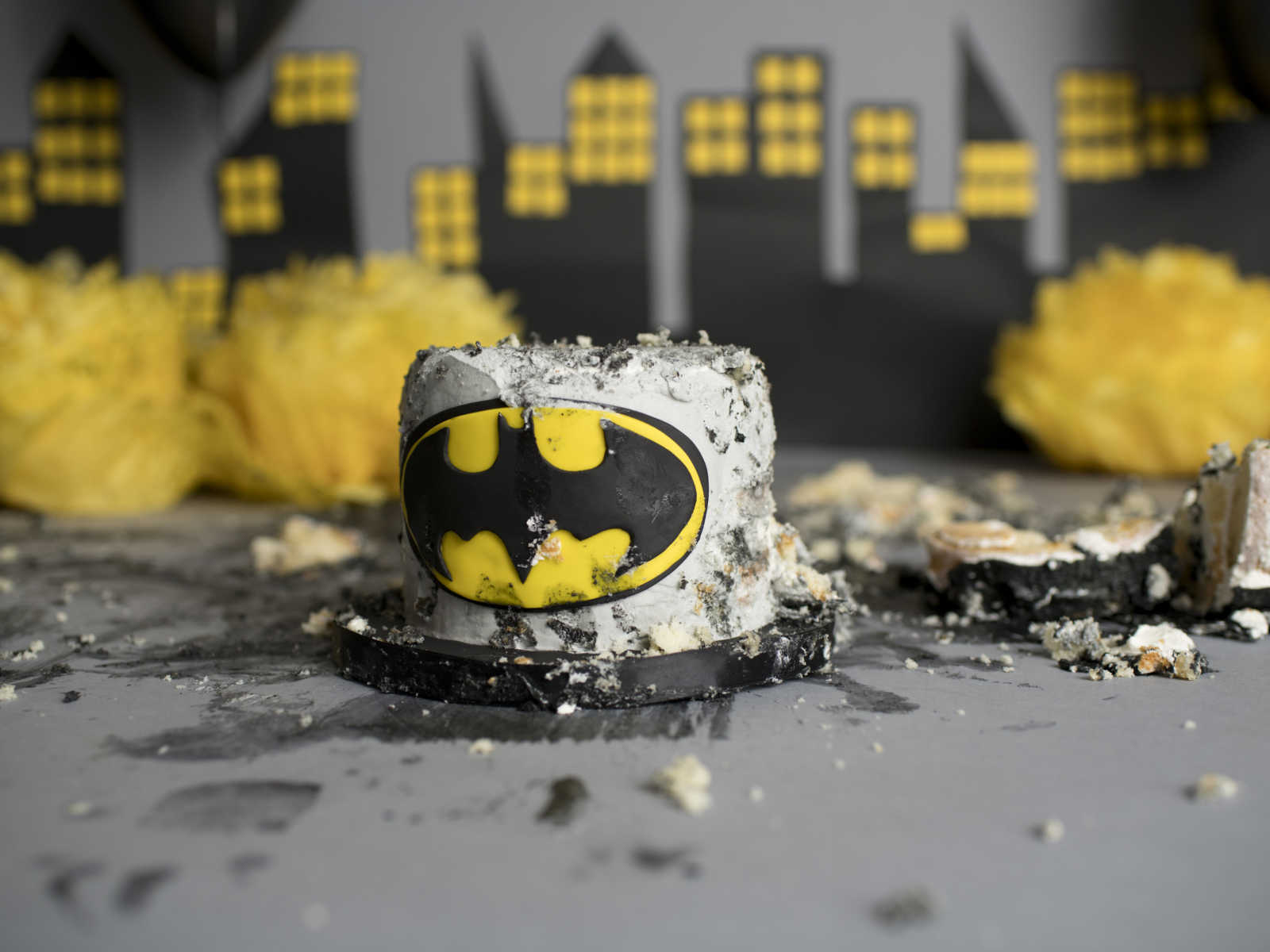 bottom tier of destroyed cake with batman logo on it in front of Gotham city backdrop