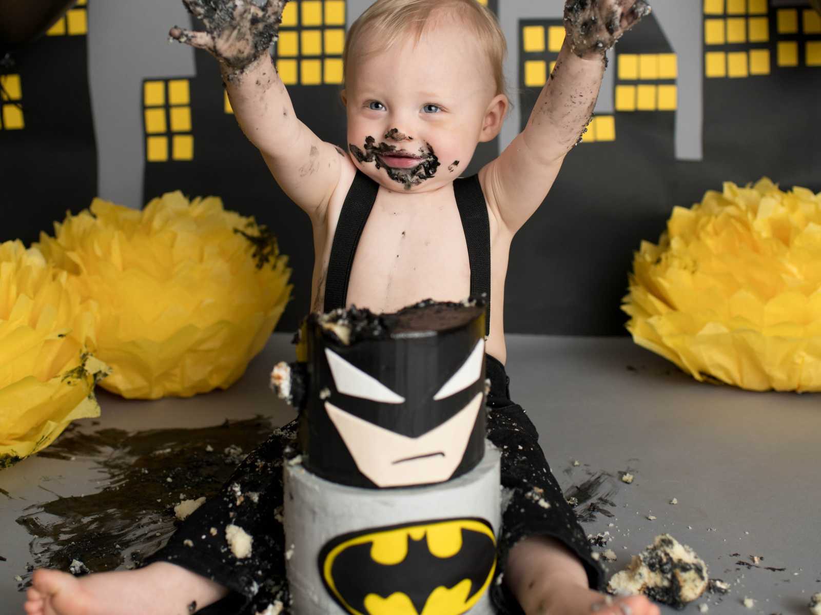 infant boy covered in black frosting raises hands in the air with batman themed cake in front of him