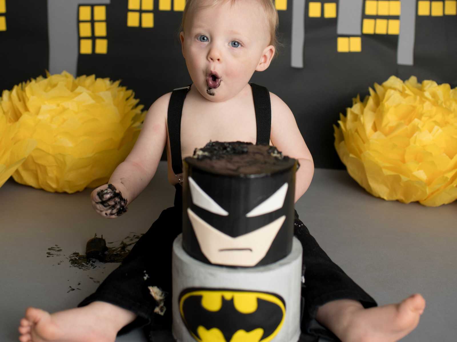 infant boy makes silly face while holding cake in his hand sitting behind batman batman cake
