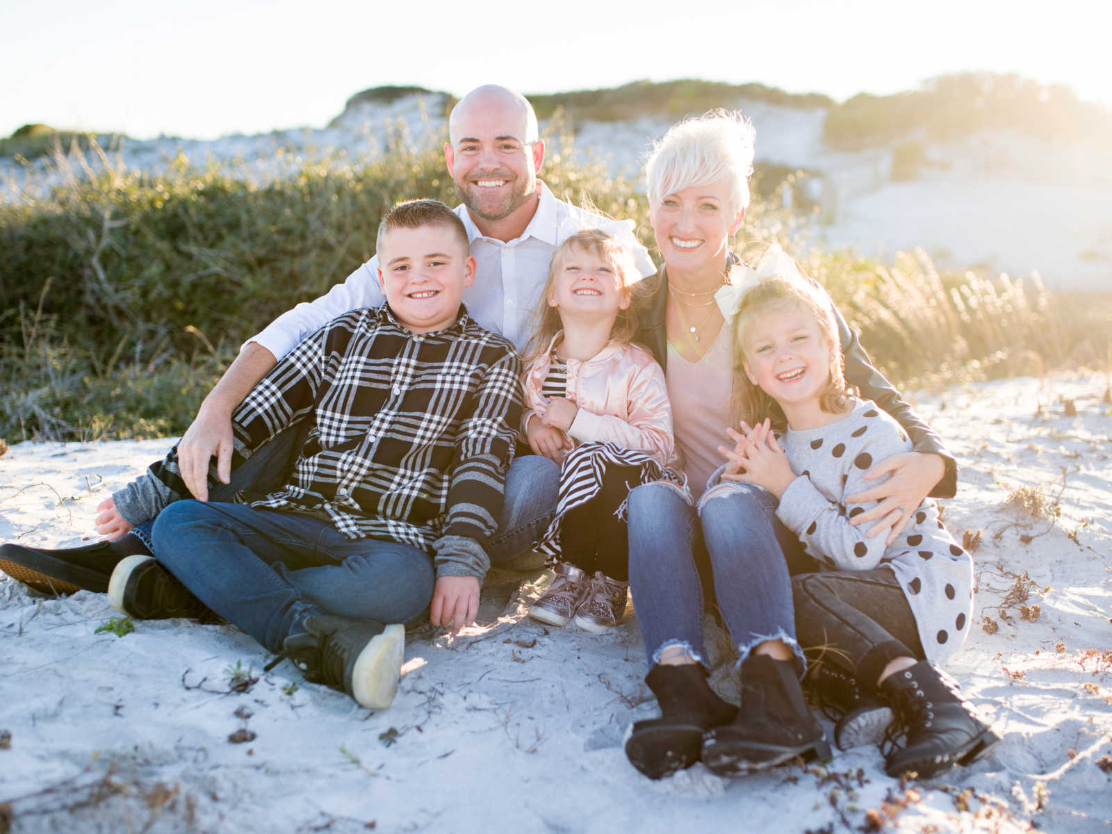 mother sits on beach with two daughters in arms next to husband and stepson