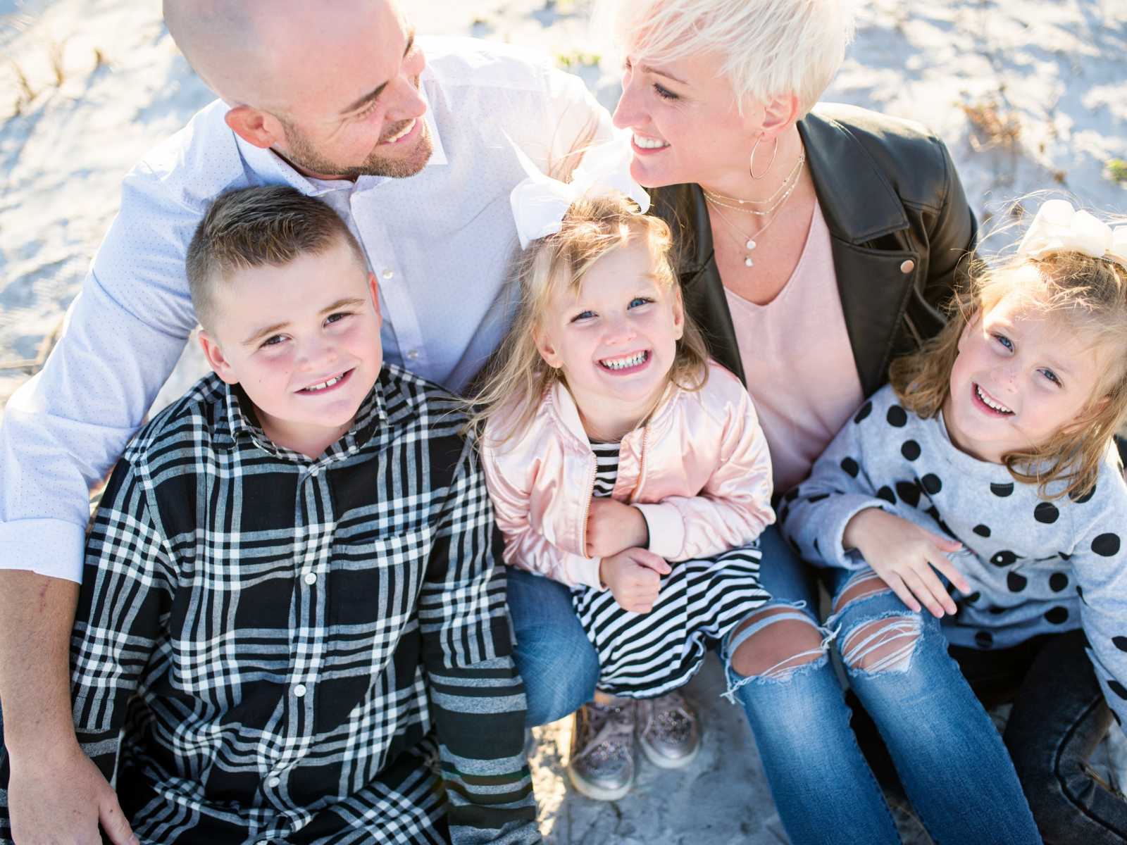 new step siblings look up smiling on parents laps on the beach while husband and wife smile at each other 