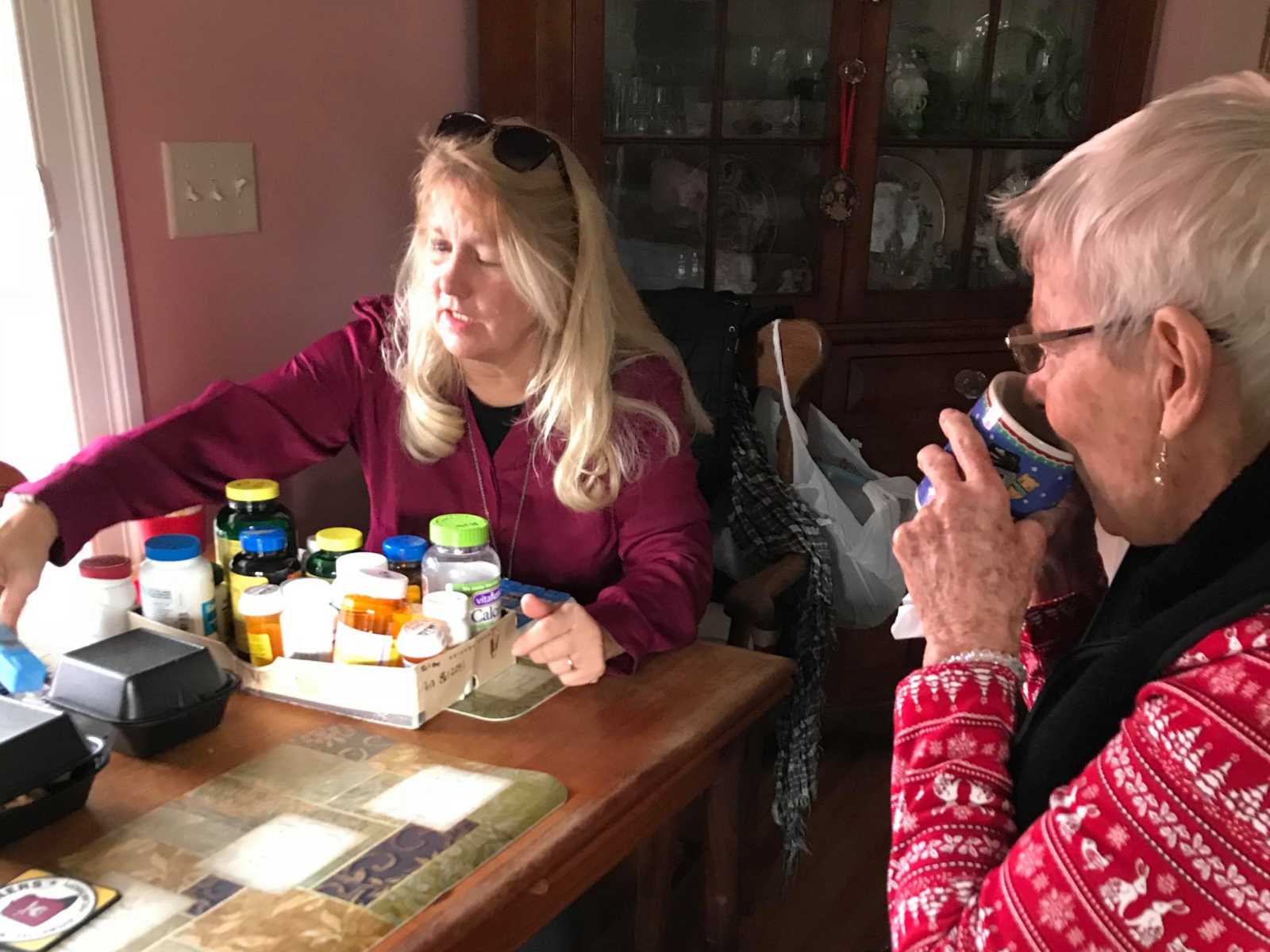 Daughter of woman with dementia sorts pill bottles while mother sips out of a mug 