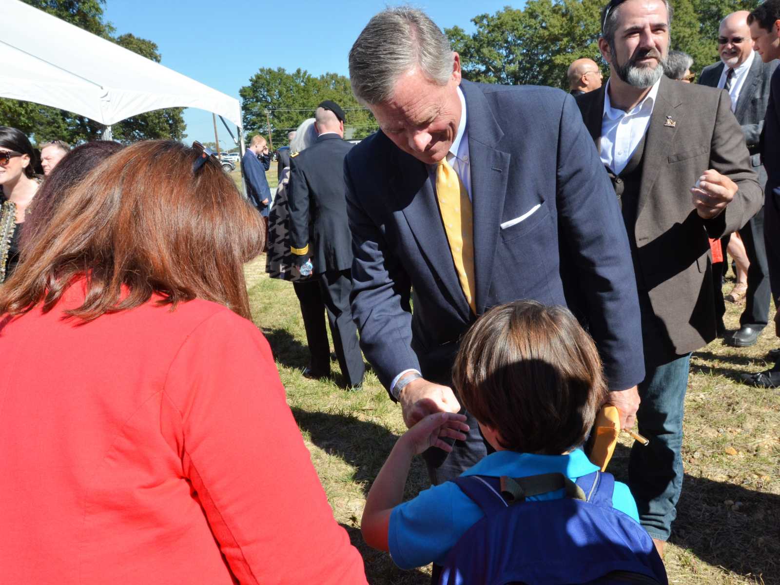 man in suit leans over to shake hand of young boy who is wearing a backpack with adults surrounding