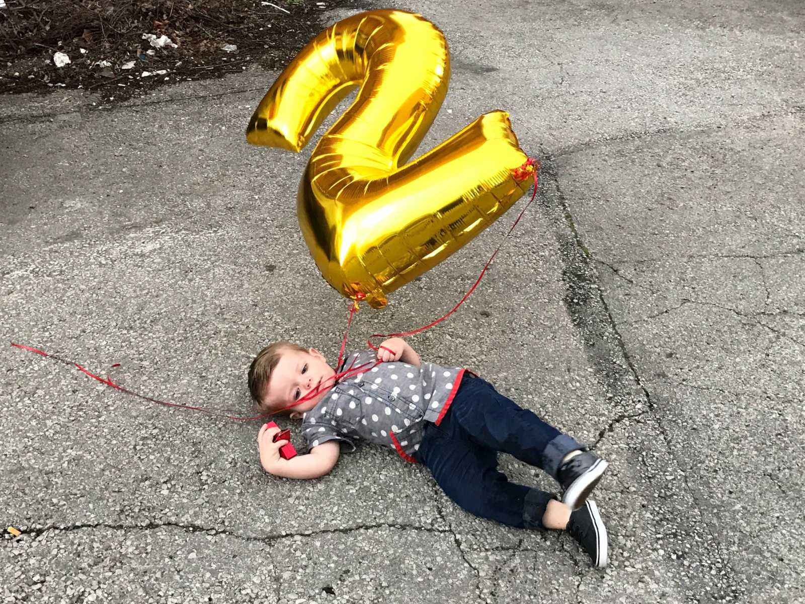 Toddler lays on his back in driveway holding on to large gold balloon that is in shape of number 2
