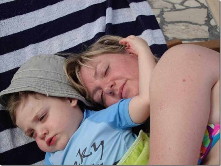 mother in bathing suit lying on striped towel on ground with arms wrapped around son with blue rash guard and bucket hat on