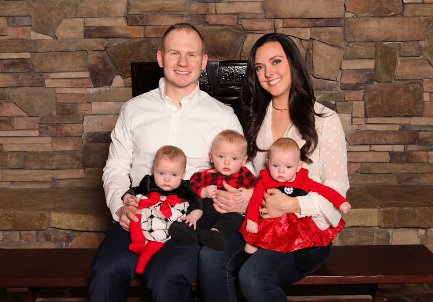 mother and father sit in front of fire place with three babies sitting on their laps