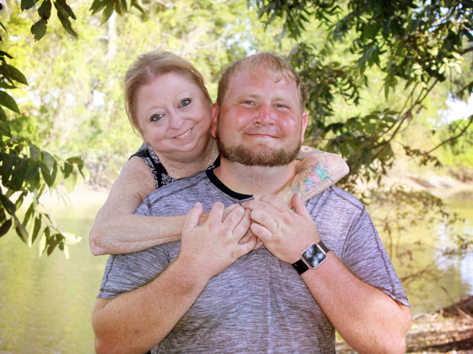 woman standing behind man with arms wrapped around him in front of body of water