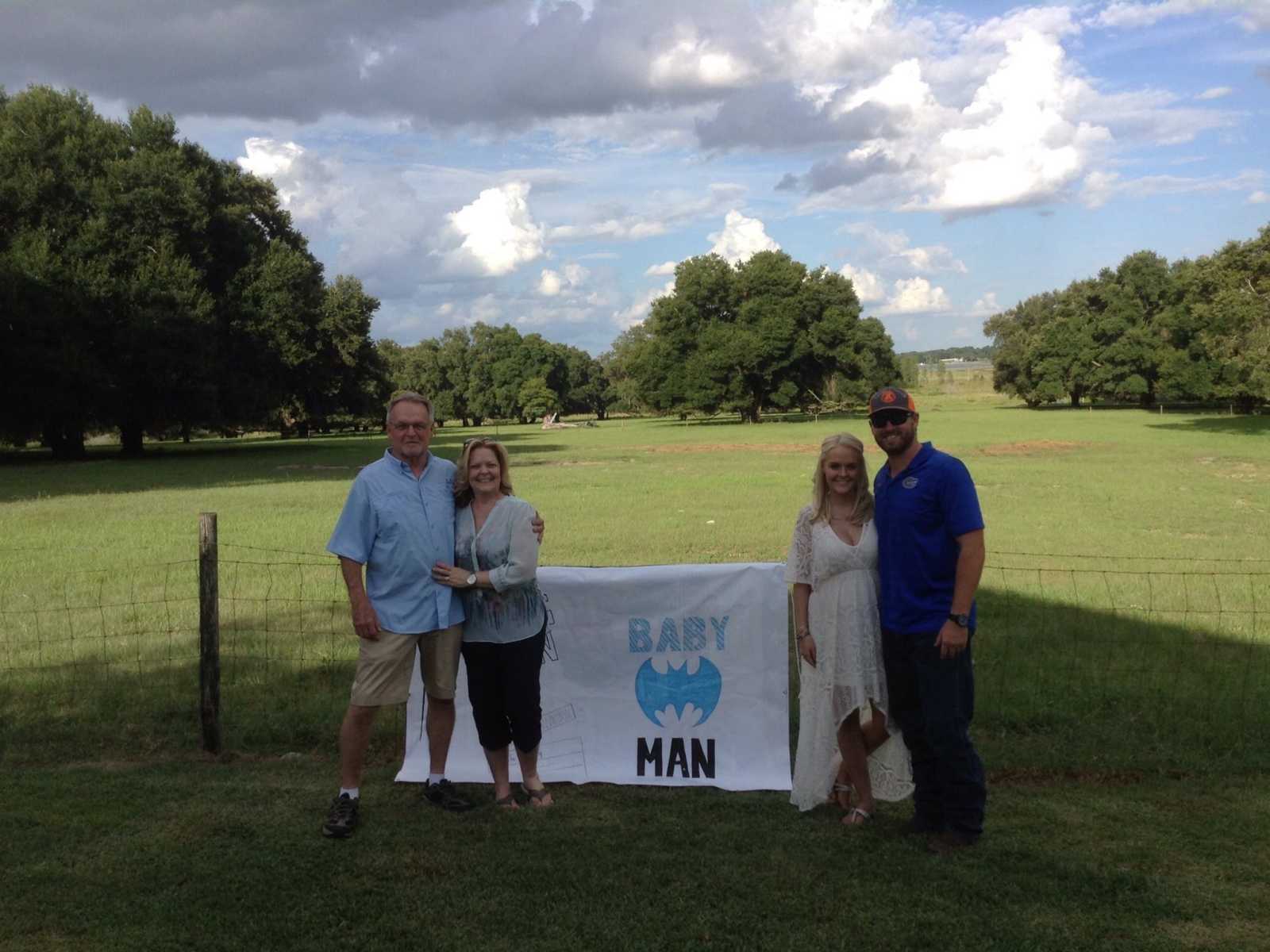 white sign hanging on wire fence that says, "baby man" with batman logo and an older and younger couple on either side