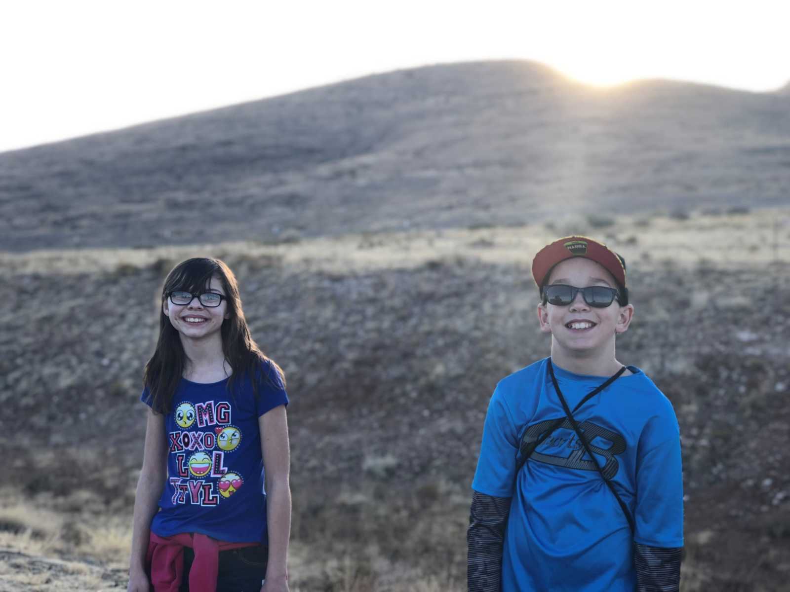 young girl in glasses smiles standing a a short distance from young boy with sunglasses on with mountains in background