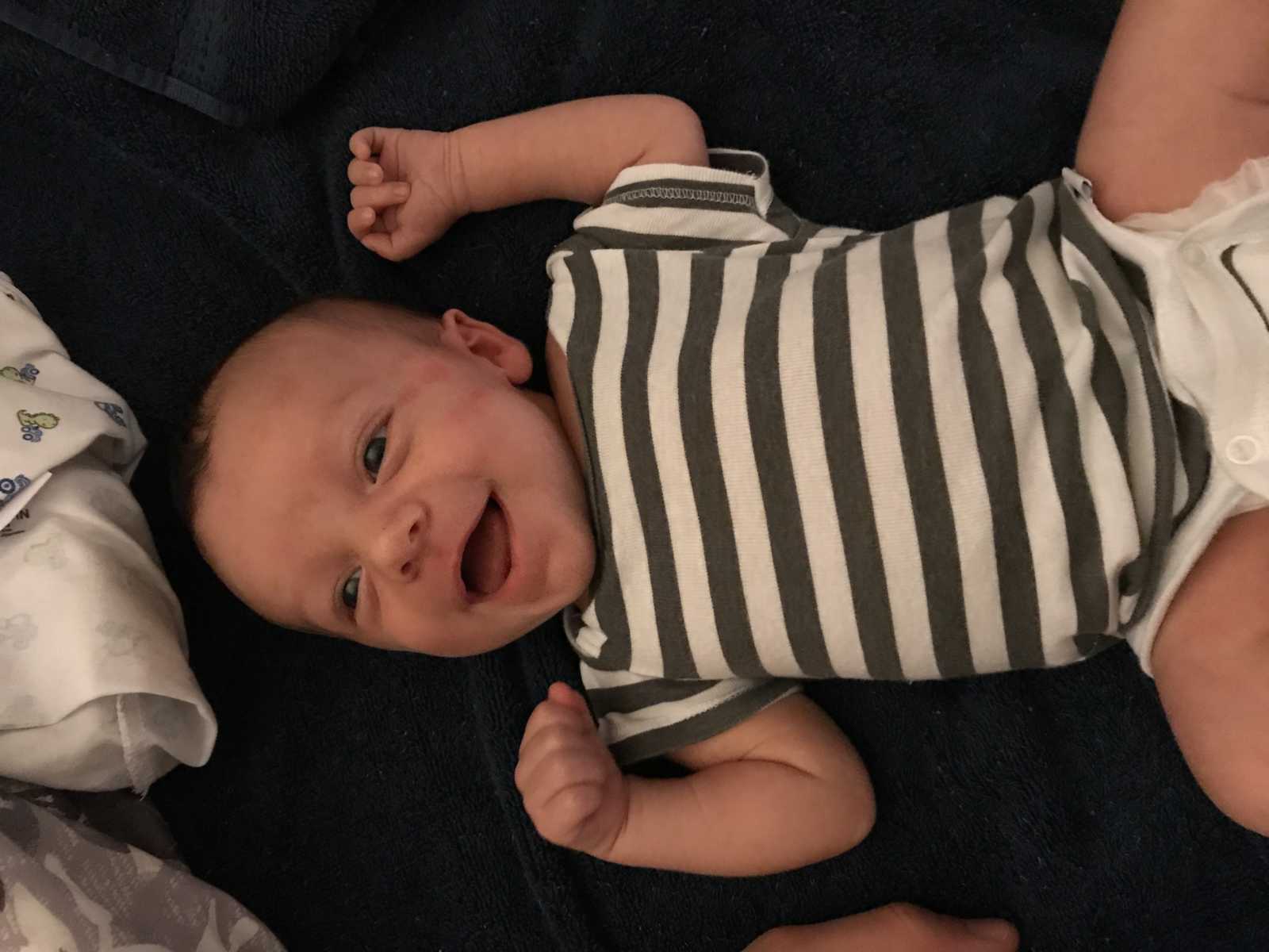 newborn wearing a green and white striped onesie smiles while lying on his back