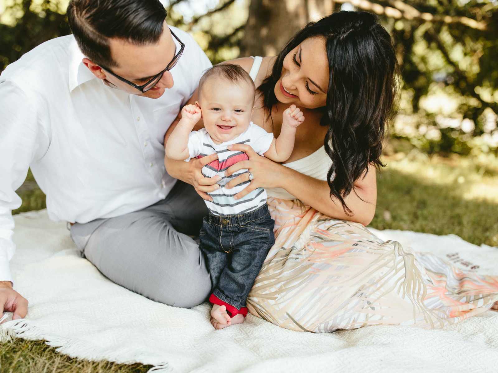 woman who struggled with postpartum sits on blanket on grass next to husband holding her son up