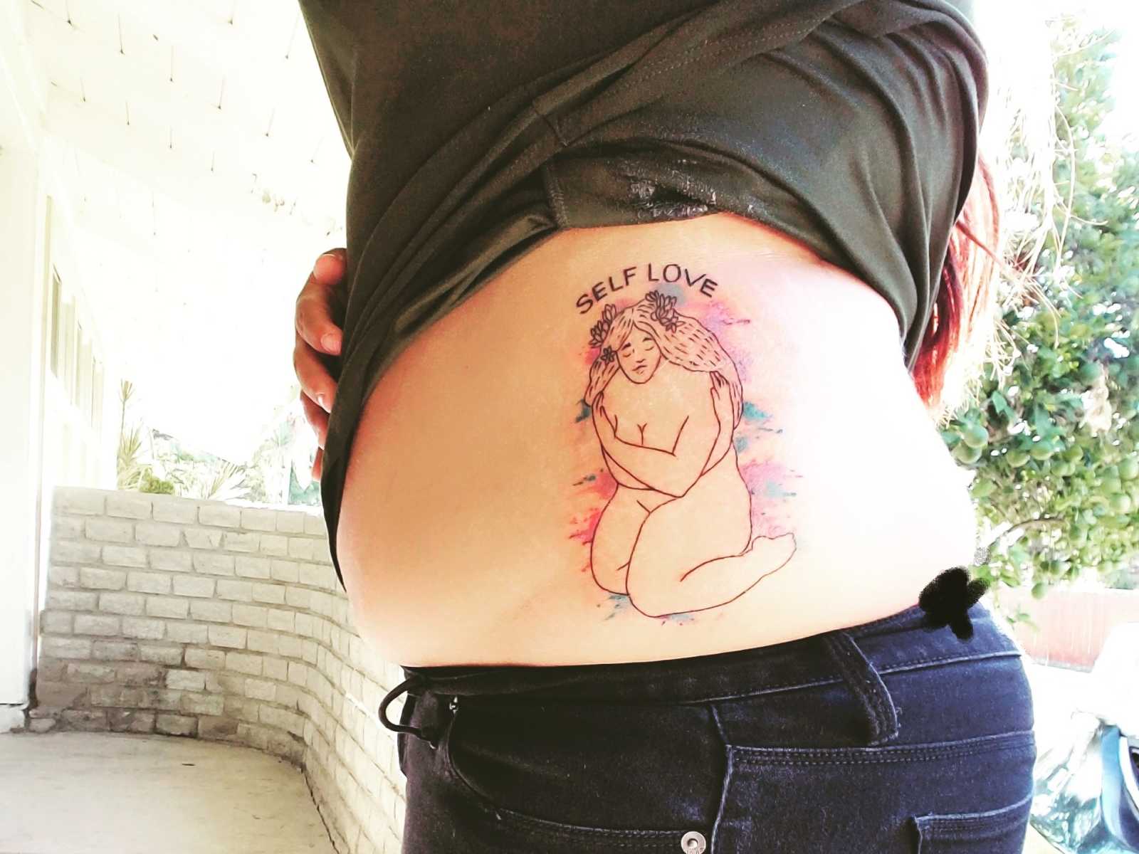 picture of woman's side with tattoo of woman sitting and holding her breasts with words "self love" arching over it