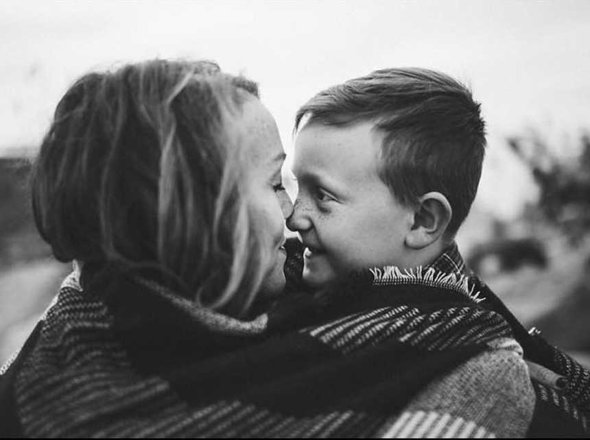 mother and son wrapped in blanket nose to nose while son looks at mother with smile