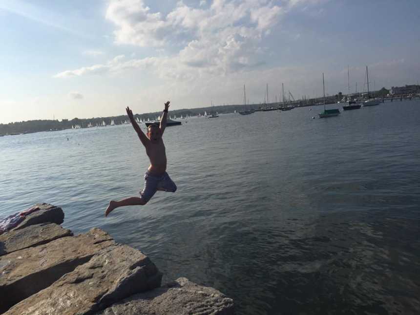 young boy jumps of rocks with hands in the air into body of water with sailboats in background