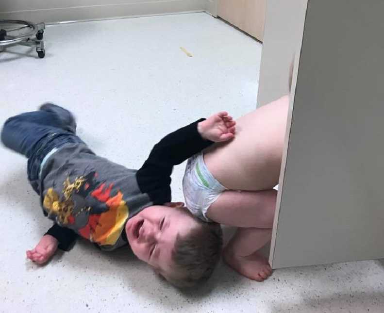 Toddler cries on floor of doctor's office next to sibling in diaper
