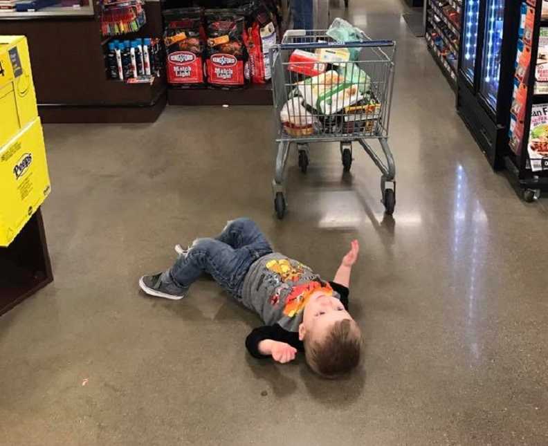 Toddler lying on the floor of grocery store next to child size shopping cart