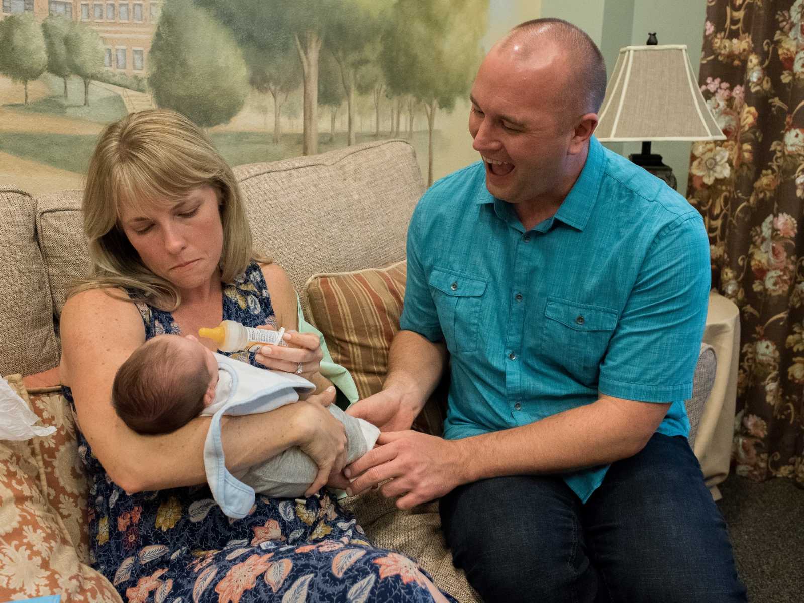 adoptive mother feeds baby with bottle on couch while adopted father looks at son with mouth open