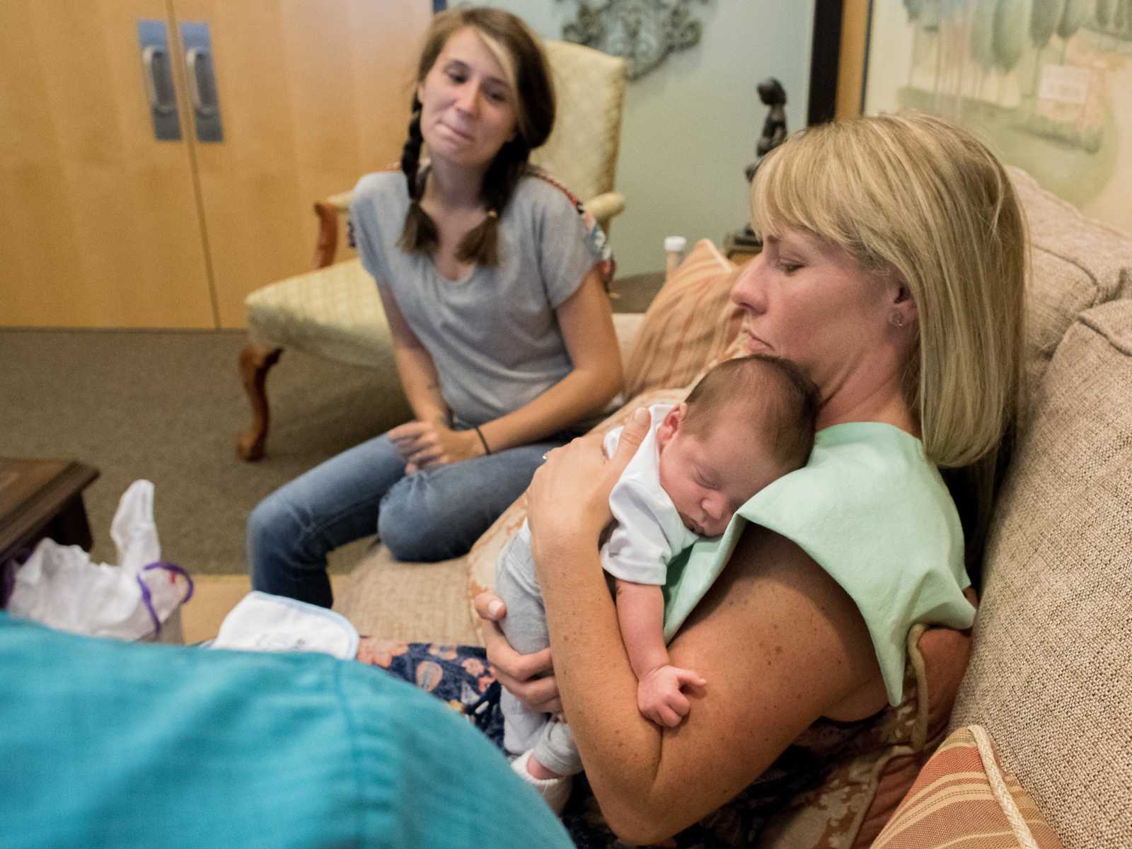 adopted mother sits on couch with baby to her chest while birth mother smiles next to her