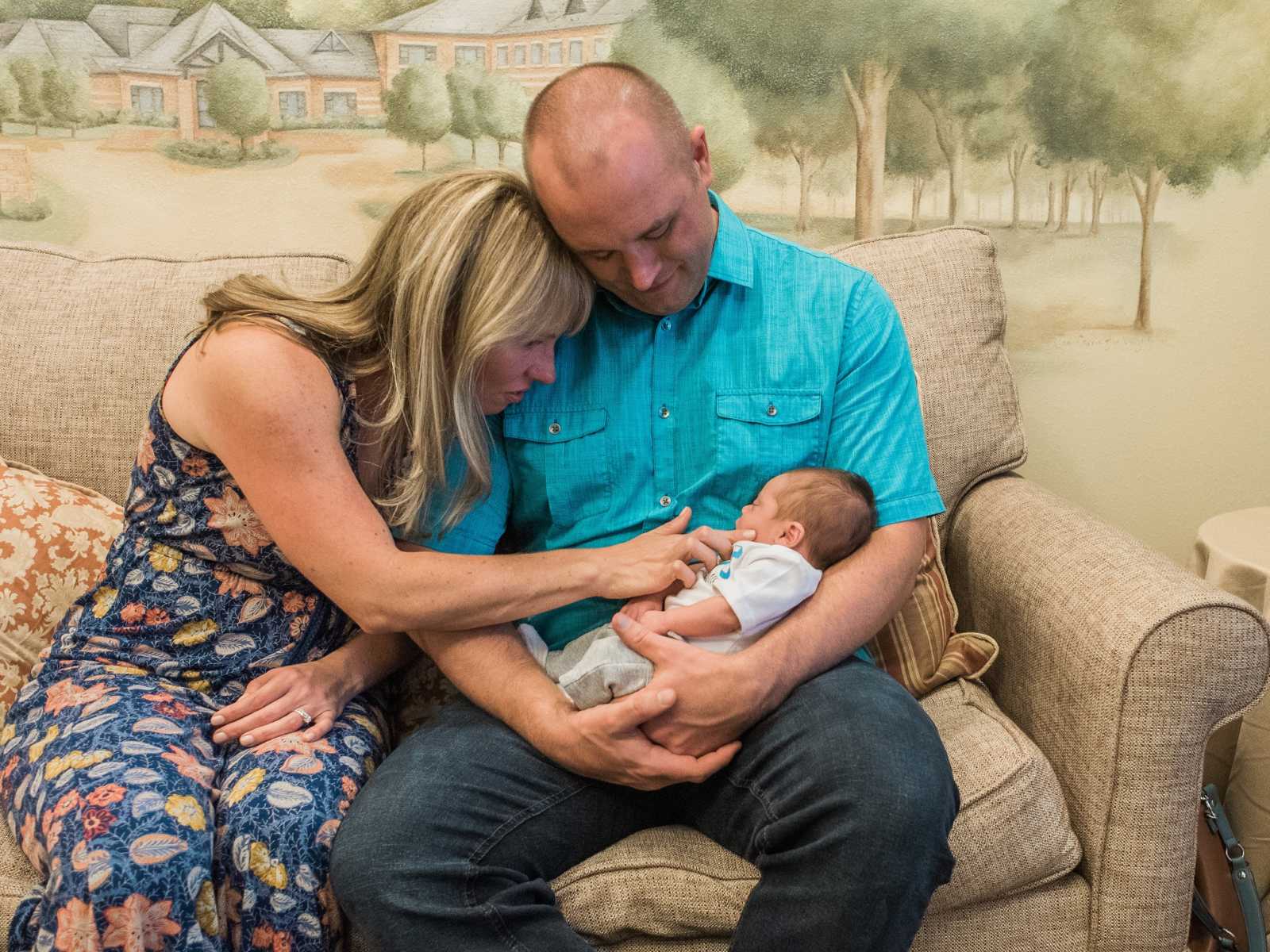 mother and father sit on couch while looking down at baby in fathers arms