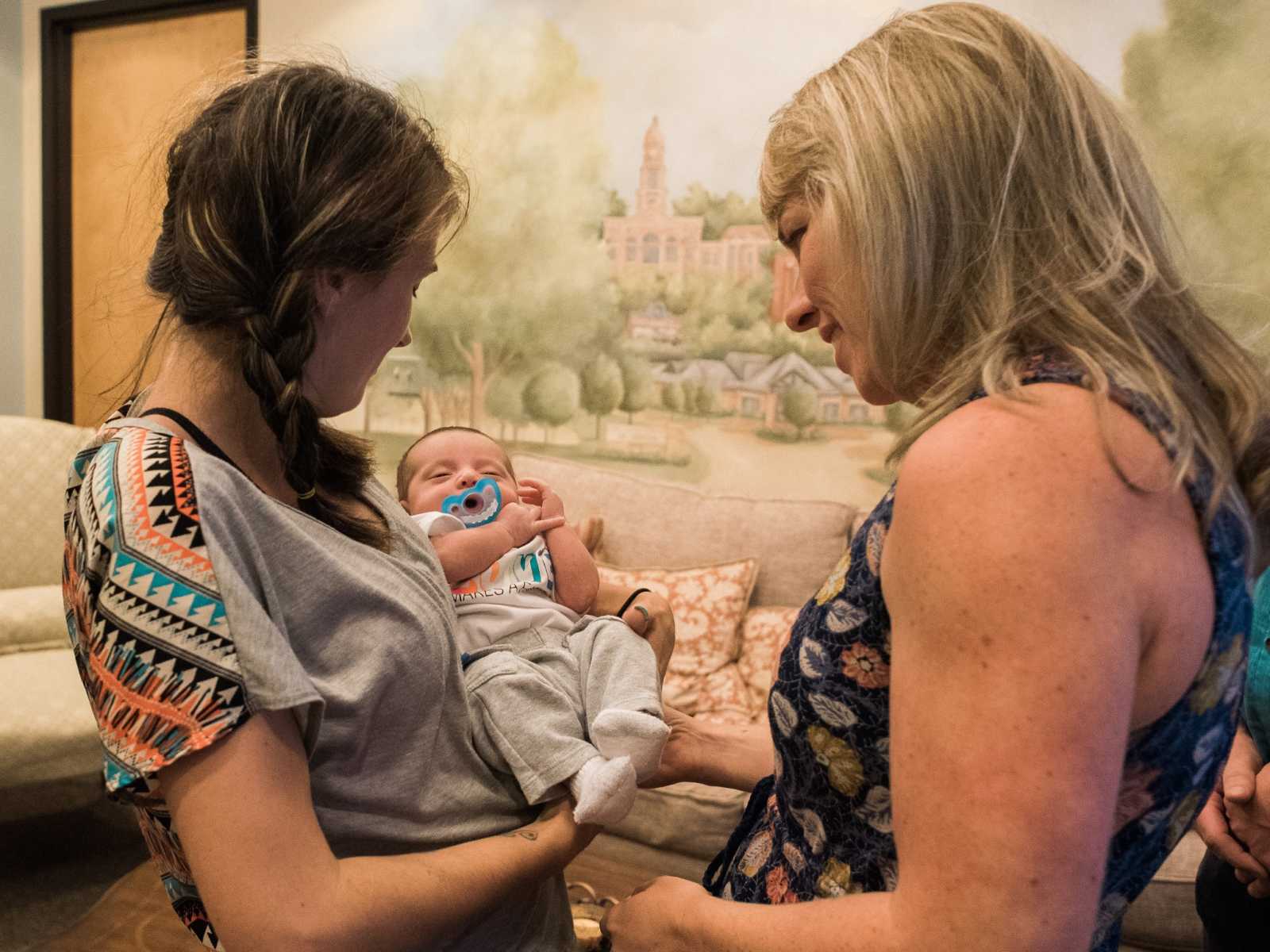 birth mother holds looks down at son in her arms next to adoptive mother