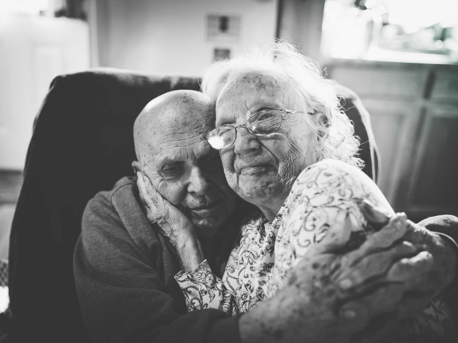 elderly wife sits on her husbands lap as he wraps his arms around her 