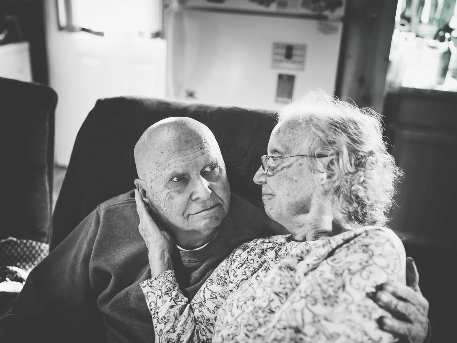 elderly wife sits on husbands lap with is arm around her