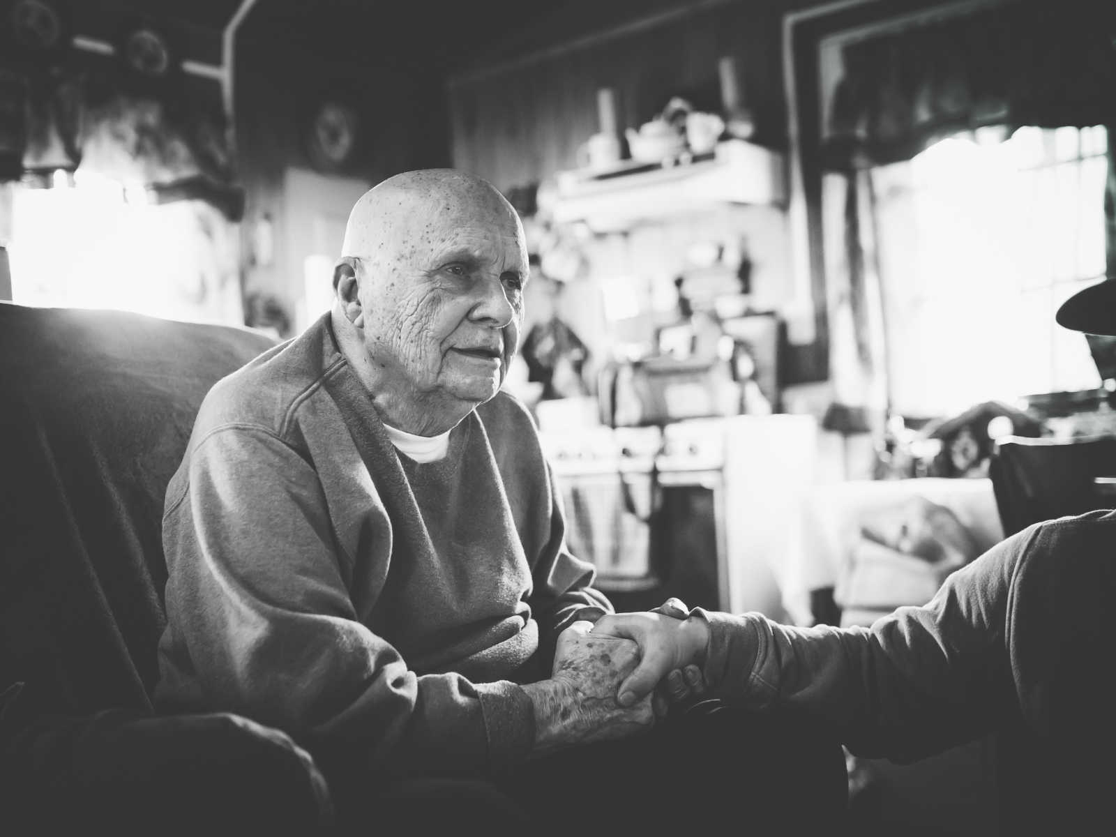 elderly man leans forward in his chair holding someones hand