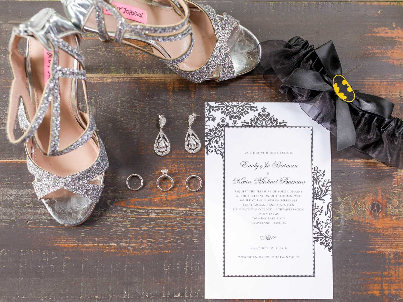 wedding invitation on wooden surface surrounded by silver earrings and rings, silver sparkly heels, and batman themed garter 