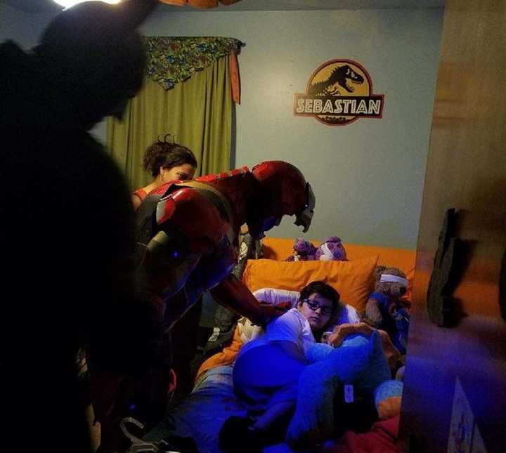 Boy with cancer lies in bed as iron man leans over him with hand on his back while batman and mother watch