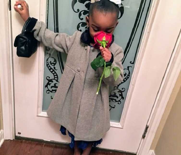 little girl stands in gray coats holding the front door knob while smelling red rose