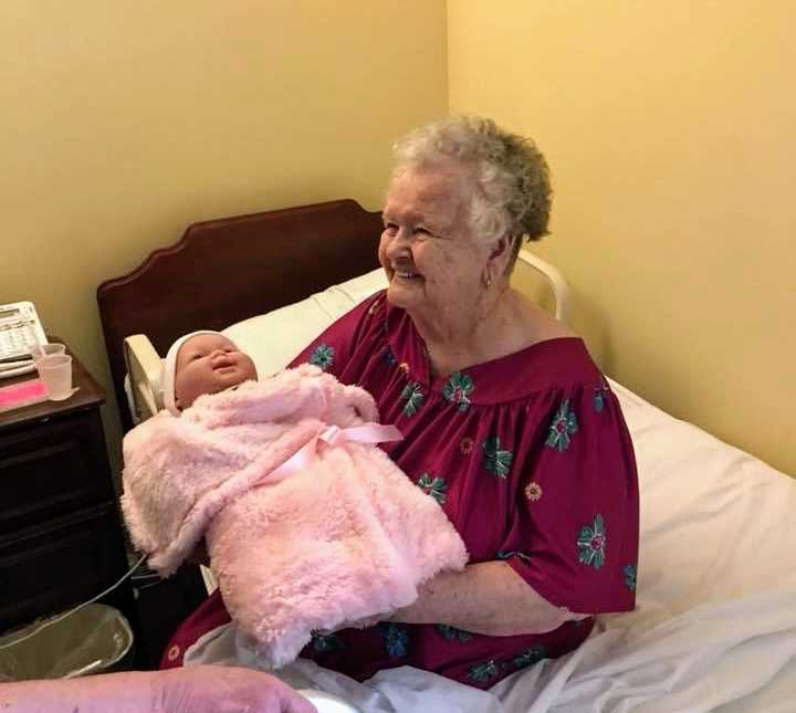 alzheimer patient sitting in bed smiling while holding baby doll wrapped in pink blanket 