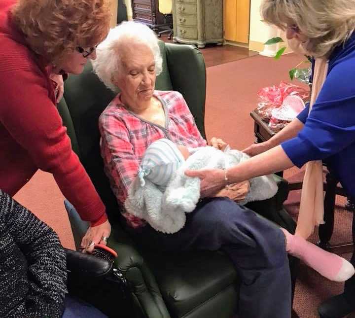 woman hands baby doll to elderly woman sitting down as another woman stand over chair of elderly woman